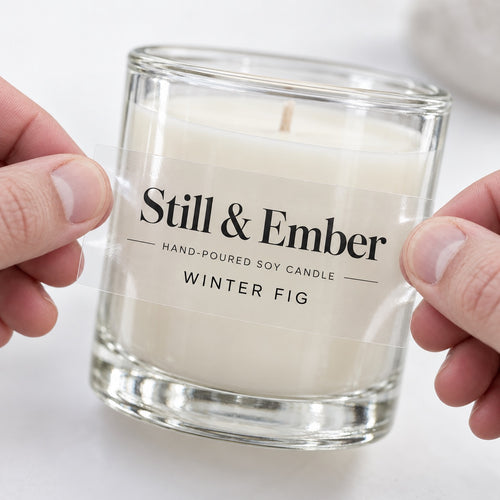Hands applying a custom clear black text label sticker to a transparent glass candle jar.