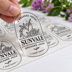 Hand holding a custom clear Sunvale orchard juice label above a sheet of matching transparent black print stickers.