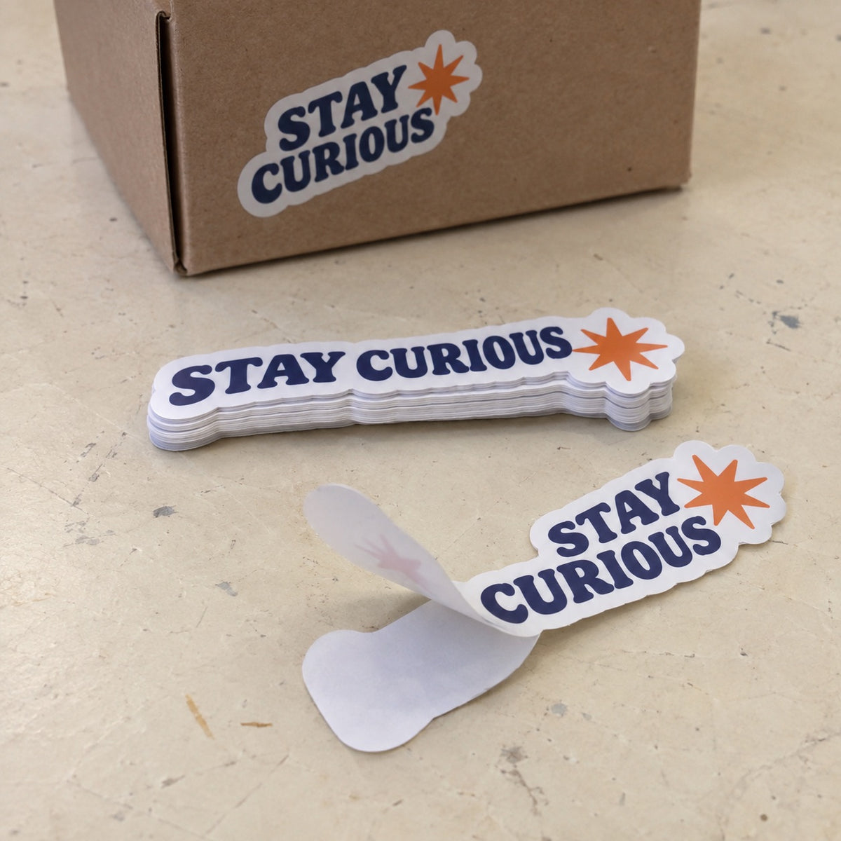 Custom Stay Curious die cut stickers shown in a stack, being peeled, and applied to a kraft shipping box.