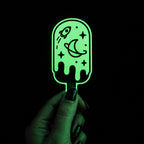 Hand holding a green glow-in-the-dark popsicle sticker featuring a rocket, moon, planet, and stars.