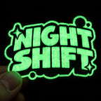 Hand holding a custom green glow-in-the-dark sticker with bold Night Shift lettering against a black background.