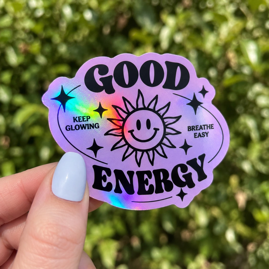 Hand holding a purple custom holographic sticker reading Good Energy with a smiling sun and rainbow shine.