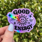Hand holding a purple custom holographic sticker reading Good Energy with a smiling sun and rainbow shine.