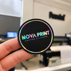 Hand holding a custom black logo sticker with holographic silver lettering and a rainbow foil border.
