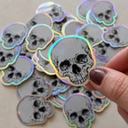 Custom die-cut holographic skull sticker held above a collection of matching silver rainbow foil stickers.