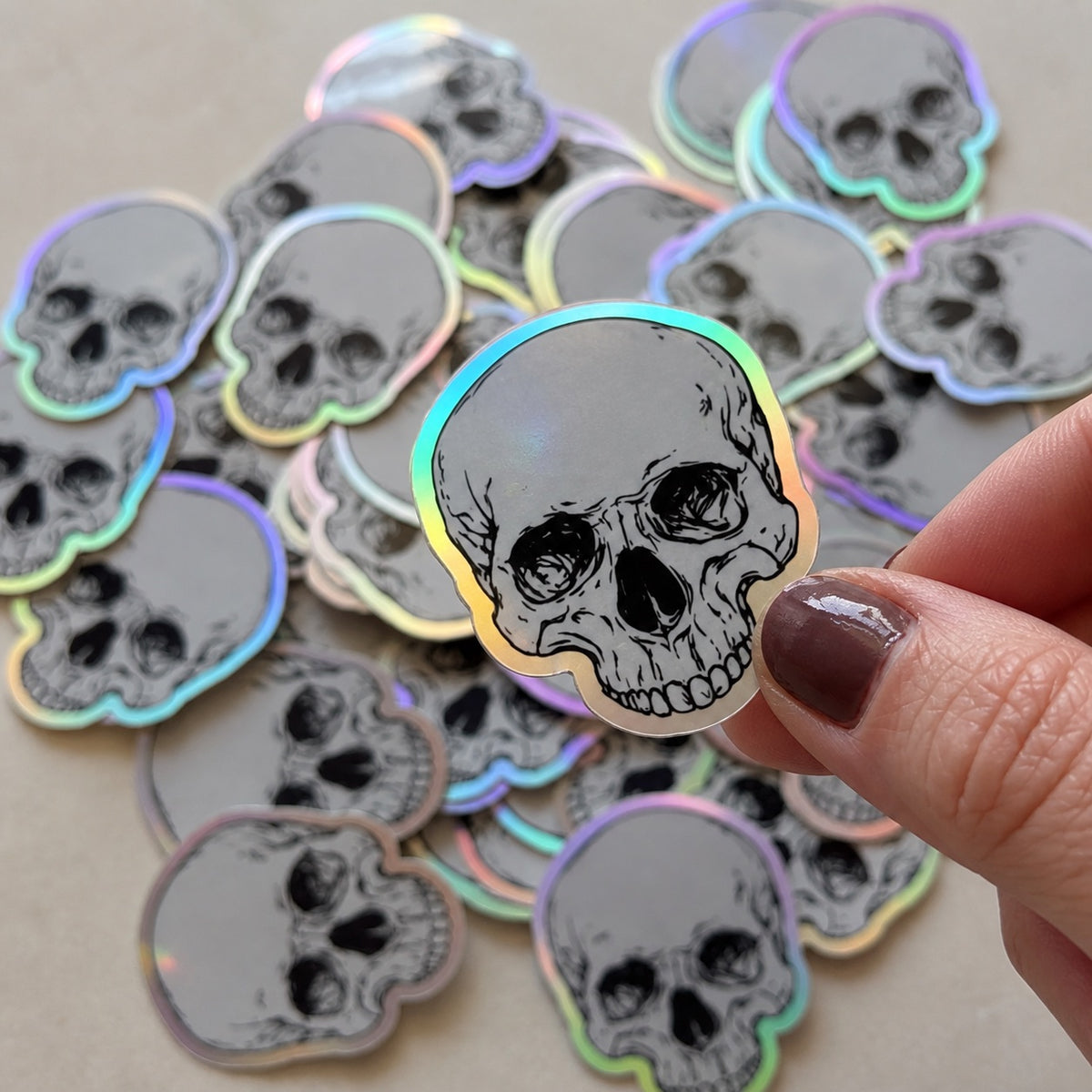 Custom die-cut holographic skull sticker held above a collection of matching silver rainbow foil stickers.