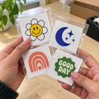Hands holding individually packaged custom kiss cut stickers with flower, moon, rainbow, and Good Day designs.